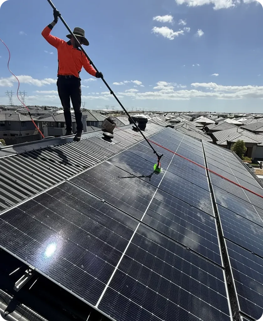 Technician cleaning a large residential rooftop solar system using a long-reach brush and purified water